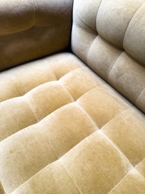 A close-up view of a plush, velvet-tufted cushion on a chair or sofa, showcasing its textured and luxurious finish in a muted golden brown color. The upholstery features a grid-like pattern with deep button tufting.