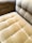A close-up view of a plush, velvet-tufted cushion on a chair or sofa, showcasing its textured and luxurious finish in a muted golden brown color. The upholstery features a grid-like pattern with deep button tufting.