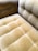 A close-up view of a plush, velvet-tufted cushion on a chair or sofa, showcasing its textured and luxurious finish in a muted golden brown color. The upholstery features a grid-like pattern with deep button tufting.