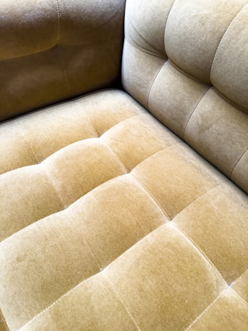 A close-up view of a plush, velvet-tufted cushion on a chair or sofa, showcasing its textured and luxurious finish in a muted golden brown color. The upholstery features a grid-like pattern with deep button tufting.