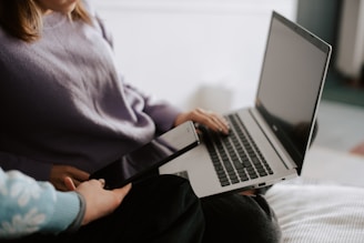 A person is sitting with a laptop on their lap, typing or navigating it while another person hands over a tablet. The setting appears to be casual and indoors, possibly at home.