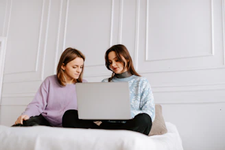two women sitting on a bed looking at a laptop
