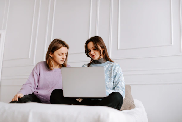 two women sitting on a bed looking at a laptop