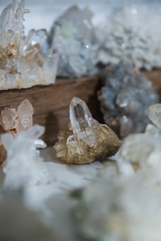Various crystals and mineral formations displayed on a wooden surface, showcasing different shapes and transparency. The focus is on a central clear crystal with a surrounding collection of similar geological specimens.