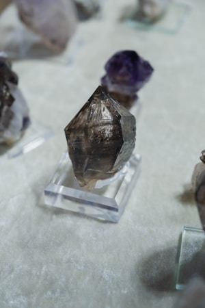 A collection of crystal-like rocks placed on transparent stands, with a prominent dark, pointed crystal in the foreground. The surface beneath is a soft, textured white fabric.
