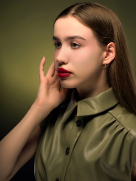 A young woman with long brown hair, wearing an olive green collared shirt, is posing with her hand gently touching her face. The background is a gradient of green shades, creating a soft, calming ambiance.