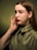 A young woman with long brown hair, wearing an olive green collared shirt, is posing with her hand gently touching her face. The background is a gradient of green shades, creating a soft, calming ambiance.