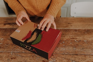 Hands opening a cardboard pizza box on a rustic wooden table. The box is decorated with Italian flag colors and a design that includes brush strokes.