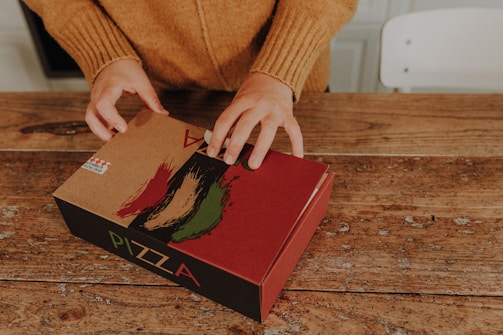 Hands opening a cardboard pizza box on a rustic wooden table. The box is decorated with Italian flag colors and a design that includes brush strokes.