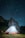 A brightly illuminated tent is set up on a grassy area under a clear night sky filled with stars. Behind the tent, there are tall, dark rock formations, adding a dramatic backdrop. The scene is tranquil and the stars are scattered across the sky, creating a sense of wonder.
