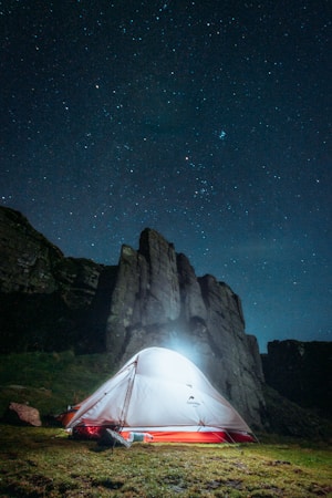 A brightly illuminated tent is set up on a grassy area under a clear night sky filled with stars. Behind the tent, there are tall, dark rock formations, adding a dramatic backdrop. The scene is tranquil and the stars are scattered across the sky, creating a sense of wonder.