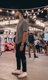 A person stands in a lively arcade setting at night, surrounded by string lights and various arcade game machines. The scene is bustling with activity as other people are visible in the background, engaging with the games or walking by. The signage reads 'Game Zone', indicating the entertainment venue.