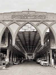 An intricately designed Islamic bazaar entrance features detailed tile work and calligraphy above a long, covered walkway. The interior is lined with arches and shops. The marketplace is bustling with people, cars, and motorcycles.