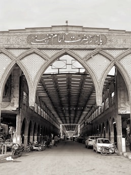 An intricately designed Islamic bazaar entrance features detailed tile work and calligraphy above a long, covered walkway. The interior is lined with arches and shops. The marketplace is bustling with people, cars, and motorcycles.