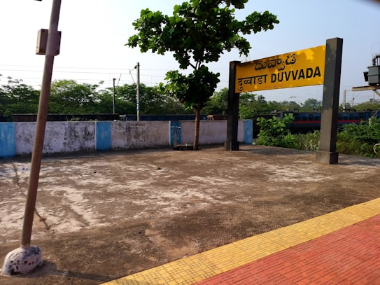 A train station platform with a sign that reads Duvvada in multiple languages. The platform is empty, with a few patches of vegetation visible. There is a tree next to the sign, casting a shadow on the concrete surface. In the background, there is a train track and a partially visible train, as well as some dense greenery and utility poles.