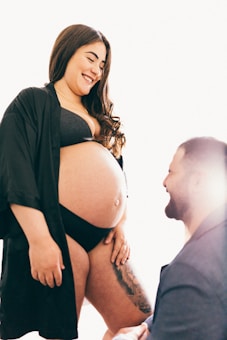 A pregnant woman is smiling warmly, wearing a black robe and undergarments. She stands in a softly lit environment, interacting with a man kneeling beside her who is partially visible. Her long hair cascades over her shoulders, and she seems to be enjoying a tender and joyful moment.