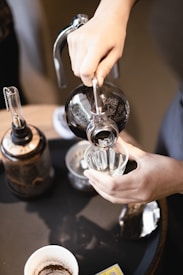 Hands carefully pour liquid from a siphon coffee maker into a glass on a table. Several coffee-making tools, including a filter and a cup with coffee residue, surround the main action. The scene captures a moment of focus and precision in a coffee brewing process.
