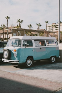 A pristine 1960s Volkswagen Transporter T1 parked on a sunlit vintage street, showcasing its classic rounded design and two-tone blue paint.