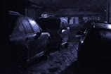 Several cars are parked in a dimly lit area behind a chain-link fence. The scene has a wet, reflective surface on the vehicles, indicating that it might have rained recently. The overall lighting is low, creating a moody atmosphere.