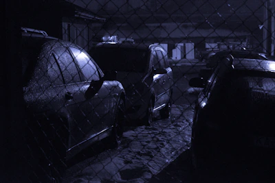 Several cars are parked in a dimly lit area behind a chain-link fence. The scene has a wet, reflective surface on the vehicles, indicating that it might have rained recently. The overall lighting is low, creating a moody atmosphere.