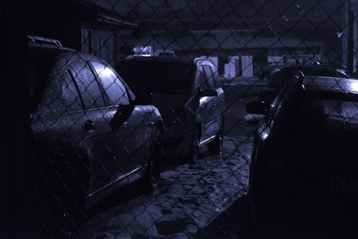 Several cars are parked in a dimly lit area behind a chain-link fence. The scene has a wet, reflective surface on the vehicles, indicating that it might have rained recently. The overall lighting is low, creating a moody atmosphere.
