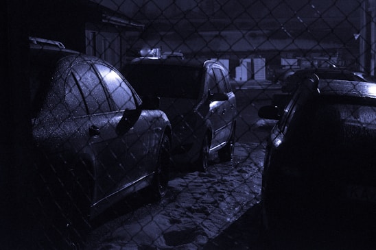 Several cars are parked in a dimly lit area behind a chain-link fence. The scene has a wet, reflective surface on the vehicles, indicating that it might have rained recently. The overall lighting is low, creating a moody atmosphere.