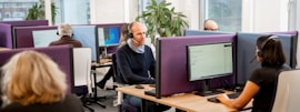 A modern office environment with several people working at computer stations. Individuals are wearing headsets, indicating a call center setting. The workspace is divided by purple privacy screens and features large desktop monitors. A green plant adds a touch of nature to the otherwise clean and professional atmosphere. Natural light comes through the windows, enhancing the bright and focused work environment.