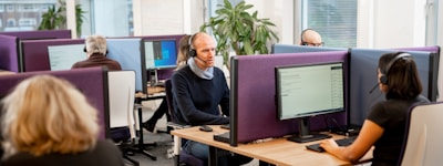 A modern office environment with several people working at computer stations. Individuals are wearing headsets, indicating a call center setting. The workspace is divided by purple privacy screens and features large desktop monitors. A green plant adds a touch of nature to the otherwise clean and professional atmosphere. Natural light comes through the windows, enhancing the bright and focused work environment.