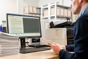 A person is seated at a desk in an office environment, holding papers while looking at a computer screen displaying a document. Stacks of papers are arranged on the desk, and a printer is situated next to the computer. Shelves filled with boxes are visible in the background.