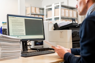 A person is seated at a desk in an office environment, holding papers while looking at a computer screen displaying a document. Stacks of papers are arranged on the desk, and a printer is situated next to the computer. Shelves filled with boxes are visible in the background.
