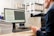 A person is seated at a desk in an office environment, holding papers while looking at a computer screen displaying a document. Stacks of papers are arranged on the desk, and a printer is situated next to the computer. Shelves filled with boxes are visible in the background.