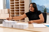 A person seated at a desk, organizing a stack of papers. Shelves filled with parcels and a potted plant in the background suggest an office or mailroom setting. The person has a focused expression, wearing a black shirt, and reaching out to pick up papers.