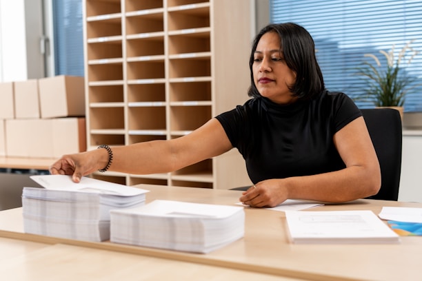 A person seated at a desk, organizing a stack of papers. Shelves filled with parcels and a potted plant in the background suggest an office or mailroom setting. The person has a focused expression, wearing a black shirt, and reaching out to pick up papers.