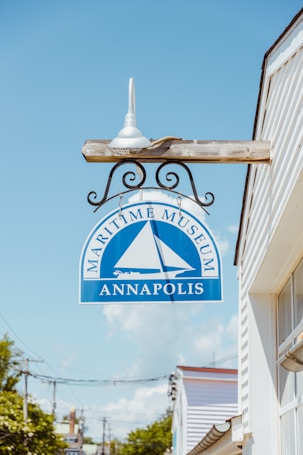 A sign for the Maritime Museum in Annapolis, featuring a blue background with a white boat graphic. The sign is mounted on a decorative metal bracket attached to a wooden pole. The pole is fixed to the side of a white building. The background shows part of a street with power lines and trees under a clear blue sky.