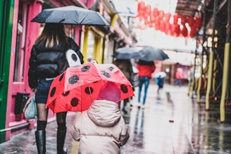 A child wearing a pink coat and hat holds a ladybug-themed umbrella with black dots. Several people walk along a wet, reflective city street, carrying black umbrellas. Vibrant colors, such as pink walls, green doors, and red lanterns, line the sides of the street.