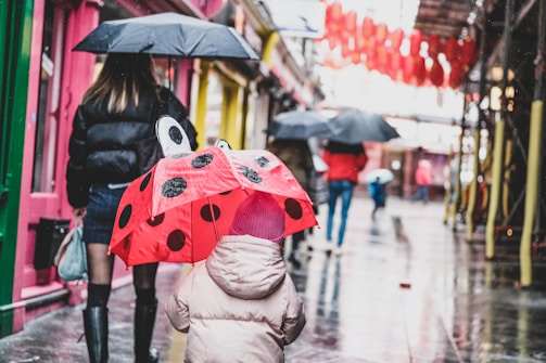 A child wearing a pink coat and hat holds a ladybug-themed umbrella with black dots. Several people walk along a wet, reflective city street, carrying black umbrellas. Vibrant colors, such as pink walls, green doors, and red lanterns, line the sides of the street.