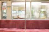 A train interior with red cushioned seats, visible signage in Japanese, and a metal pole. The window view shows a blurred exterior with a passing train.