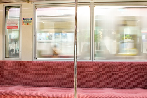 A train interior with red cushioned seats, visible signage in Japanese, and a metal pole. The window view shows a blurred exterior with a passing train.