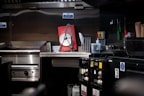 A commercial kitchen with stainless steel countertops and kitchen equipment, including a griddle and a dish rack holding a large red and white sign. Various kitchen tools and appliances are visible, such as knives in a holder, a bottle of cleaning liquid, and switches on the wall.