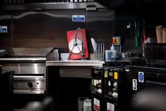 A commercial kitchen with stainless steel countertops and kitchen equipment, including a griddle and a dish rack holding a large red and white sign. Various kitchen tools and appliances are visible, such as knives in a holder, a bottle of cleaning liquid, and switches on the wall.