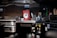 A commercial kitchen with stainless steel countertops and kitchen equipment, including a griddle and a dish rack holding a large red and white sign. Various kitchen tools and appliances are visible, such as knives in a holder, a bottle of cleaning liquid, and switches on the wall.