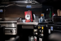 A commercial kitchen with stainless steel countertops and kitchen equipment, including a griddle and a dish rack holding a large red and white sign. Various kitchen tools and appliances are visible, such as knives in a holder, a bottle of cleaning liquid, and switches on the wall.