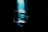 A clear glass bottle with a spray nozzle is placed against a dark background with a vibrant, blue light illuminating its backside, creating a striking reflection and shadow effect.