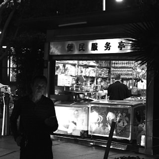 A black and white image of a small convenience store or kiosk. Inside, a person stands behind the counter surrounded by a variety of products, including snacks and drinks. The store front is brightly lit, contrasting with the darkness outside. Another person is visible in the foreground, partially in shadow, holding an object that could be a phone.