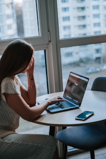 A woman sits at a round table near a large window, using a laptop and looking at the screen. The room appears to have a modern design with natural light coming through the window. A smartphone is placed on the table beside her, and outside, there are blurred views of apartment buildings.