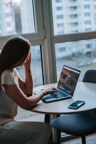 A woman sits at a round table near a large window, using a laptop and looking at the screen. The room appears to have a modern design with natural light coming through the window. A smartphone is placed on the table beside her, and outside, there are blurred views of apartment buildings.