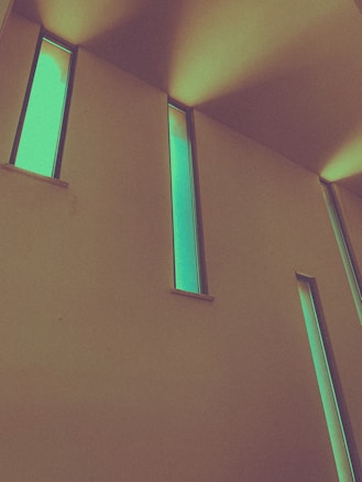 Tall, narrow windows with a glowing green hue are set against a muted wall, creating an abstract and modern architectural design. Light and shadows play dramatically across the ceiling and walls.