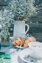 A peaceful outdoor setting featuring a table adorned with delicate blue flowers in glass jars and a ceramic pitcher. Fresh croissants rest on a white plate beside a cup of tea, creating a serene breakfast or brunch atmosphere.