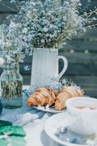 A peaceful outdoor setting featuring a table adorned with delicate blue flowers in glass jars and a ceramic pitcher. Fresh croissants rest on a white plate beside a cup of tea, creating a serene breakfast or brunch atmosphere.