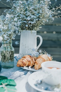 A peaceful outdoor setting featuring a table adorned with delicate blue flowers in glass jars and a ceramic pitcher. Fresh croissants rest on a white plate beside a cup of tea, creating a serene breakfast or brunch atmosphere.